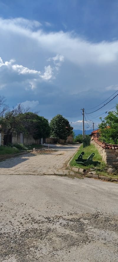 The image captures a sunny, tranquil street scene in Beguntsi, Bulgaria, viewed along a path or road. The upper half of the frame is dominated by a bright blue sky adorned with large, fluffy white clouds.

On the right side of the image, an unpaved, rough dirt road with scattered gravel extends into the distance. Adjacent to it, on the left, is a paved path constructed from rectangular, light-colored bricks, some of which appear weathered. Between this brick path and a low stone wall, a strip of vibrant green grass is visible. Resting on this grassy strip are two distinct, dark green, angular metal objects with cut-out designs, possibly decorative elements or abstract outdoor furniture.

Behind the stone wall, which is topped with traditional reddish roof tiles, are glimpses of houses with similar red-tiled roofs, partially obscured by various trees. The trees vary in density, with some showcasing lush green foliage. A tall utility pole with overhead wires stands prominently in the mid-ground, extending towards the distant horizon. Faint outlines of mountains can be seen beneath the expansive sky in the far background. Several small, dark specks, likely birds, are visible flying high in the sky.

The scene depicts a quiet, rural or semi-rural environment under bright daylight. No people are present, and there is no visible text.