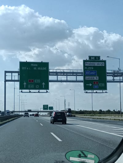 This media file captures a perspective from inside a vehicle traveling on a highway. The scene is dominated by a bright, slightly overcast sky with large, puffy white clouds. The highway itself is multi-lane, with visible lane markings and other vehicles ahead and to the sides.
Two prominent green highway signs are visible, directing traffic. The left sign indicates routes to "PRAHA" (Prague), "ÚSTÍ N. L." (Ústí nad Labem), and "HR. KRÁLOVÉ" (Hradec Králové), with directions and road numbers like E55 and E65. The right sign points towards "Pražský okruh" (Prague Ring Road), "PLZEŇ" (Plzeň), "ŘÍČANY", and "Letiště" (Airport), along with road numbers 0, E50, and 101. The number 12 is also visible on this sign.
The presence of Czech place names and road numbering systems (like E55, E50) strongly suggests the location is in the Czech Republic, specifically near Říčany as indicated by the sign. The time of day appears to be daytime, given the lighting conditions. The weather is fair with significant cloud cover. The overall mood is one of travel and navigation. The reflection on the lower right suggests a car interior, possibly a dashboard or windshield. No people are distinctly visible.