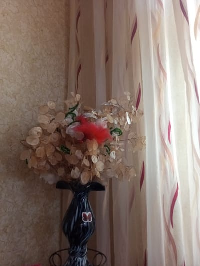 This is a forensic, matter-of-fact description of a still life image likely taken in a residential setting in Chernyshevskoye, Russia. The scene features a decorative arrangement of dried flowers and artificial foliage in a black and white swirled vase. The vase has a white butterfly decal on its front. Behind the vase and floral arrangement are sheer curtains with a pattern of thin, elongated burgundy and peach-colored leaf-like shapes. The wall to the left of the arrangement has a textured, light brown finish. No people or actions are depicted in the image.