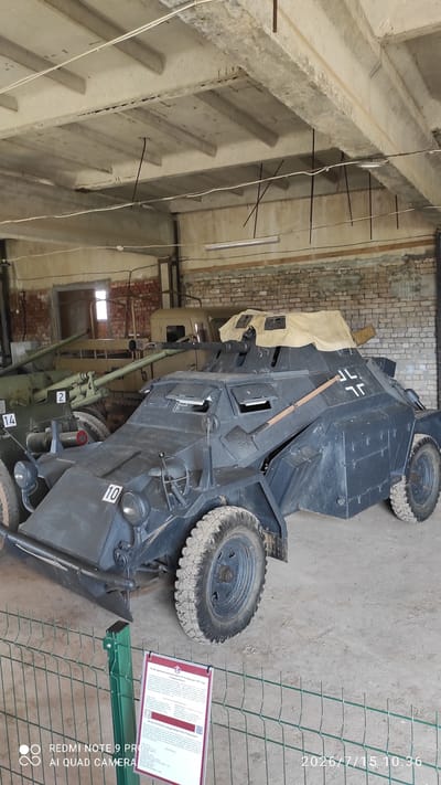 A dark grey armored car with a canvas top is parked indoors on a dirt floor. The vehicle has a license plate with the number 10 on the front. There are also German cross symbols on the side of the vehicle. Behind the armored car, there is a military cannon with the number 2 and 14 visible. Another tan colored vehicle is visible further back. The scene appears to be inside a museum or exhibition hall, with exposed concrete beams and brick walls. A informational sign is placed in front of a green fence, with text in Russian and English, which identifies the armored car as a "Sd.Kfz. 222 armored car" and the cannon as a "45-mm tank gun Model 1937 'Baby Fedor'". The location is identified as Laszanski village, Belarus.