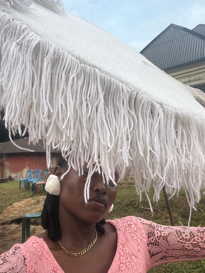 A close-up, portrait-style photograph, rotated 90 degrees counter-clockwise, depicts a dark-skinned person, likely a woman, outdoors in Aghalokpe, Nigeria. The upper left portion of the frame is dominated by a large, dense arrangement of white fringes, resembling thick yarn or thread, which partially obscures the person's face, covering her forehead and left eye.

The person wears a pink lace top or dress with an intricate, open-work pattern. Her hair appears to be styled in dark braids, and a white, rose-shaped decorative element is visible near her right ear. A gold chain necklace adorns her neck. Her expression is neutral, and her gaze is directed slightly downwards.

The background elements are mostly out of focus. In the upper left, a corrugated metal roof is visible beneath a bright, overcast sky. Below the person's chin and to the right, green grass and foliage are present. Further back, colorful plastic chairs (blue, green, red) and a reddish-brown wall or building are indistinctly visible. The scene is set during the daytime, under soft, diffused lighting. No text is visible in the image.