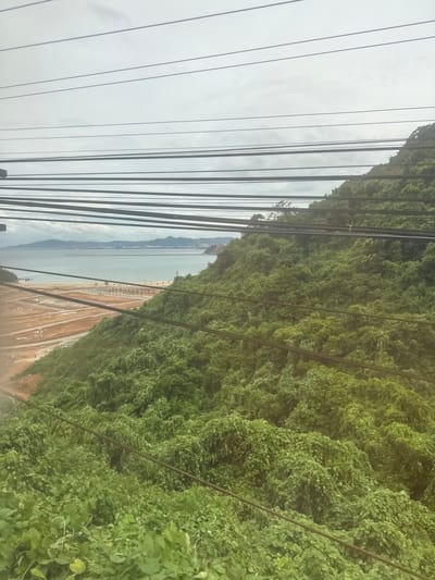 The image depicts a view from an elevated position, likely a hillside, overlooking a coastal area. Multiple thick black electrical cables stretch horizontally across the frame, obscuring parts of the landscape. Below the cables, a lush green, densely vegetated hillside slopes downwards towards a beach. The beach itself shows signs of development, with cleared areas of reddish-brown earth and visible construction stakes or foundations. Beyond the beach, a body of calm water, likely the sea, stretches to the horizon. In the distance, a hazy cityscape with buildings and mountains is visible. The sky is overcast with a pale, cloudy appearance. The scene suggests a partially developed coastal region in Hải Vân Ward, Vietnam, characterized by natural greenery, ongoing construction, and electrical infrastructure.