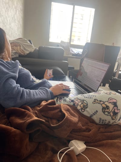 In Skhirat, Morocco, an indoor scene depicts an individual seated, partially obscured from the rear and side, engaged with electronic devices. The person, appearing to be female, wears a blue fleece top and has dark hair. A ring is visible on the fourth finger of her left hand. She operates a silver-toned laptop with her right hand on an external mouse, while holding a smartphone in her left hand, which displays a female face, indicative of a video call or image. The laptop screen shows what appears to be a spreadsheet application.
The individual is covered by a brown, textured blanket, underneath which a light blue pillow adorned with cartoon characters, including Smurfette and a character with "#GRUMPY FACE!", is visible, supporting the laptop. A white power adapter with a cable rests on the brown blanket in the foreground.
The environment includes a dark brown or black leather sofa or armchair behind the person. A large window with white frames and horizontal bars dominates the background, admitting significant natural light, partially overexposing the exterior view, which suggests another building. A white shopping bag labeled "PRESS" is situated near the window. The wall to the left features a textured, light-colored finish. Additional furniture, including other dark chairs, is partially visible in the background.