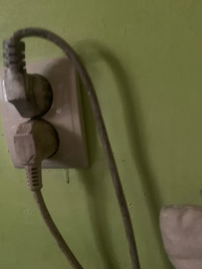 The image shows a close-up view of two electrical plugs inserted into a wall socket against a textured, light green wall. One plug is positioned above the other. Both plugs appear to be grey and have a corrugated section where the cable meets the plug. The cable from the upper plug curves upwards and out of frame to the left. The cable from the lower plug curves downwards and to the left. A shadow of the cable from the upper plug is visible on the wall. The image does not provide any information about the city of Skarydzew, Poland, nor does it depict any people or interactions. The scene appears to be indoors.