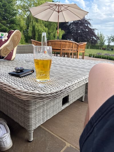 A low-angle shot captures a casual outdoor setting. A partially consumed beer and a wallet with keys rest on a light grey wicker table. A pair of sneakers, one maroon and one white, sit on the table and the patio, respectively. In the background, wooden chairs and an open umbrella are visible, set against a backdrop of green trees and a bright sky. The edge of a person's leg and clothing enters the frame from the top.