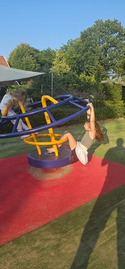 Two young girls are playing on a round playground merry-go-round in a park. The merry-go-round has a blue frame with yellow accents. One girl, with long blonde hair, is positioned on the left side of the merry-go-round, wearing a white top and patterned pants. The second girl, with long brown hair, is on the right side, wearing a green tank top and a white skirt. She is holding onto the blue frame and is suspended in the air. The merry-go-round is situated on a red rubberized surface, with green artificial turf surrounding it. In the background, there are lush green trees and bushes, and a hint of a building with a red roof is visible on the left. The lighting suggests it is late afternoon or early evening, with long shadows cast on the ground. The park appears to be located in the city of Groenlo, Netherlands.