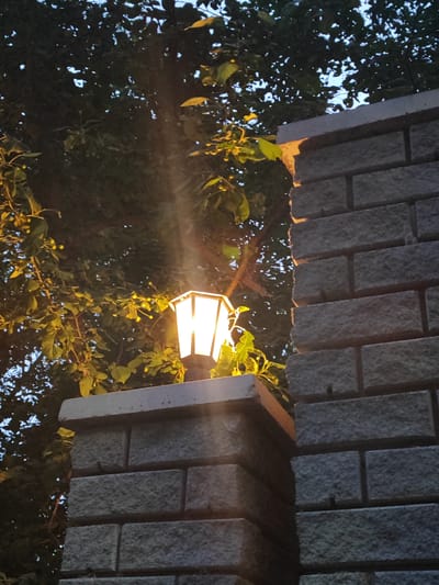 The image shows an outdoor scene at dusk, with a street lamp illuminating a section of a brick wall and nearby foliage. The street lamp, octagonal in shape with frosted glass panels, is mounted on top of a pillar constructed from light-colored, rough-hewn bricks. The lamp emits a warm, yellow light, casting rays that create a visible beam through the air. To the left of the lamp and pillar, dark green leaves and branches of a tree are visible, some of which are illuminated by the lamp. The background is a dark sky, partially obscured by the tree canopy. The brick wall to the right of the pillar is also constructed from similar rough-hewn bricks, laid in a staggered pattern. The overall lighting suggests it is either early morning or late evening. There are no people or animals visible in the image. The scene appears to be a residential or public outdoor space, possibly a garden or pathway. The specific location in the city of Slavgorod, Russia, cannot be determined from the visual information present in the image.