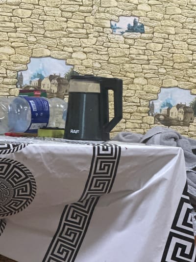The image shows a close-up view of a table covered with a white tablecloth featuring a black geometric pattern. On the table, there is a dark gray electric kettle with a silver accent and the brand name "RAF" in white letters. To the left of the kettle are several plastic water bottles and a package of what appears to be tea. A yellow sponge and a red object, possibly a pen or marker, are also on the table. In the background, a wall is decorated with a faux stone pattern and mural-like images depicting a European village scene with buildings and a bridge. The overall setting appears to be indoors, likely a kitchen or dining area. There is no discernible indication of the location being in Corlăteni, Moldova, as this information is not present in the visual cues of the image.