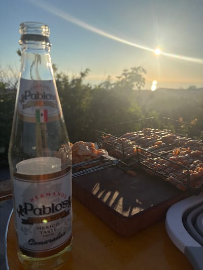 A close-up, eye-level shot captures a bottle of Pablo's Light Beer and a grill basket filled with raw shrimp, bathed in the warm glow of a sunset. The beer bottle, mostly empty, stands on a wooden surface in the foreground, its label clearly visible, featuring "HERMANOS Pablo's LIGHT BEER MEXICO TASTE FROM Camaradas" and a small Mexican flag. Behind the bottle, a grill basket holds a generous portion of uncooked shrimp, arranged in rows. The background reveals a blurred landscape of lush green foliage and the shimmering surface of the sea, with the sun setting on the horizon, casting long shadows and lens flare across the scene. The overall impression is one of a serene outdoor gathering, likely in a coastal area near Povarovka, Russia, where the evening air is filled with the promise of a grilled seafood meal.