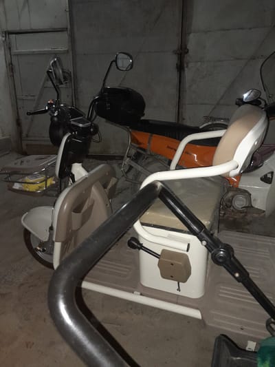 The image shows a three-wheeled electric scooter parked indoors. The scooter is predominantly white and beige, with an orange seat and accents on the rear. It features a front basket containing a yellow object. The scooter is parked on a concrete floor in what appears to be a garage or storage area, indicated by the metallic rolling door in the background. The lighting is dim, suggesting it might be evening or a poorly lit indoor space. No people are present in the image. The location is identified as Mekhrabad (Proletarsk), Tajikistan.