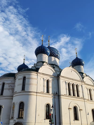 This media file is a photograph of the Assumption Cathedral in Selivanovo, Russia. The cathedral is a white building with several blue domes adorned with gold stars. The sky is a clear blue with scattered white clouds, suggesting it is daytime with fair weather. The sun is casting shadows on the building, indicating it is likely morning or afternoon.
The cathedral's architecture features multiple arched windows and ornate detailing. The prominent blue domes are topped with golden crosses, typical of Orthodox Christian churches. The scene is peaceful and majestic, highlighting the beauty of the religious structure against the backdrop of a bright sky. There are no people visible in the image, nor any specific activity taking place. The focus is solely on the architectural splendor of the cathedral. There is no visible text in the image.