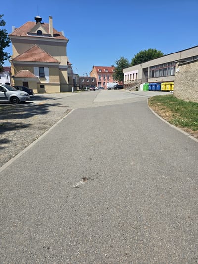 The image depicts an outdoor scene on a clear day, likely in Kostelec na Hané, Czechia, showing a pathway in the foreground with buildings and vehicles in the background. A paved pathway curves from the foreground towards the right, bordered by a gravel area on the left and grass on the right. On the left side, a light-colored car is parked in the gravel lot. Further back, a distinct yellow building with a red tiled roof stands prominently. Across the street, a modern building with a flat roof and large windows is visible, with a row of colored recycling bins (green, blue, and yellow) lined up in front of it. Several vehicles, including a white van and a dark-colored car, are parked in the vicinity of the buildings. The sky is a clear, bright blue. The scene appears to be a public area, possibly near a school or municipal building, given the presence of recycling bins.