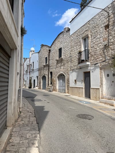 The image depicts a deserted street in Ceglie Messapica, Italy, under a clear blue sky with sparse white clouds. The asphalt road, appearing to ascend slightly, is flanked by buildings on both sides.
On the left, the immediate foreground features a light-colored building with a large, closed metal roll-down security shutter covering its ground-level entrance. Further along this side, multiple white-painted structures with visible air conditioning units and various windows and doors line the street. Rooftops in the distance display television antennas and a satellite dish.
The right side of the street is lined with buildings primarily constructed of light-colored stone with white upper sections and trim. A prominent building features a small, black wrought-iron balcony on the first floor, containing a potted plant. Below the balcony, the ground floor has a white facade with a light brown wainscoting, an address number "15," and a doorway protected by a metal chain-link screen. Adjacent to this, a white wooden door with small square window inserts is visible. Further along this side, multiple buildings feature distinctive arched doorways, some with small sets of stone steps leading up to them. These doors are typically gray or metallic in appearance. Windows on these stone buildings are dark, suggesting closed shutters.
The street surface includes a manhole cover near the center and a blue disabled parking symbol painted on the right sidewalk. Yellow painted lines are visible on the curb near the right sidewalk. A narrow, uneven cobblestone sidewalk borders the left side of the asphalt road. Strong sunlight from an elevated source casts distinct shadows of the buildings and their features onto the street and sidewalks. No individuals or vehicles are present. Minimal vegetation, including a small plant growing from the side of the leftmost building and the plant on the balcony, is visible.