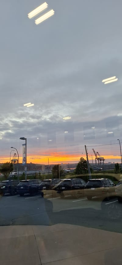 The image, taken at the Halifax waterfront, Canada, captures a sunset view reflecting on a glass surface. The sky is a canvas of orange and yellow hues, streaked with grey clouds. In the foreground, a row of dark-colored SUVs is parked in a lot. Their reflections are clearly visible on the glass.
Further in the background, industrial cranes dominate the waterfront skyline, suggesting a busy port. A street lamp and a tall banner are also visible, adding to the urban landscape. The overall scene depicts a twilight moment at a Canadian port city, with the natural beauty of the sunset contrasted against the industrial elements.