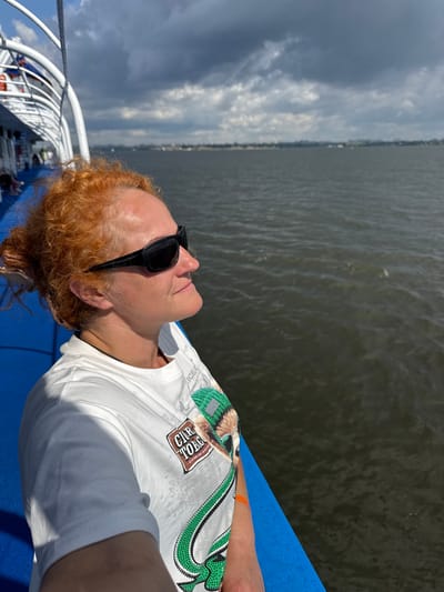 A woman with red, curly hair pulled back and wearing sunglasses is leaning on a blue railing on the deck of a boat. She is wearing a white t-shirt with a graphic print on it. The boat is moving on a body of dark water, and the sky is partly cloudy. In the distance, there is a hazy shoreline with buildings, suggesting an urban or suburban area. The location is the city of Voronovka, Russia, specifically on a body of water, likely a river or bay.