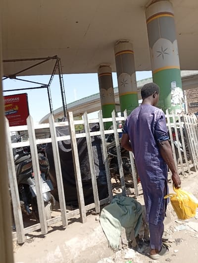The image depicts an outdoor urban scene in Tudun Wazurchi, Nigeria, under an elevated concrete structure. A single adult male, viewed from behind and the right side, stands near a light-colored metal bar fence. He wears a dark blue or purple long-sleeved tunic with subtle patterns over matching trousers and light-colored open-toed sandals. In his right hand, he holds a yellow plastic bag. His left arm is bent at the elbow.

The overhead concrete structure is supported by at least three cylindrical pillars visible in the mid-ground. These pillars are painted with alternating bands of grey, thin brown, light grey with white floral/star patterns, thin brown, and green at the base. Behind the fence, several motorcycles or scooters are parked, some partially covered by black tarpaulins. A red billboard with the text "MUSKMAN" and "BOOM premium BODY SPRAY" is partially visible on the far left. The ground consists of dirt and scattered debris, including a light green/khaki fabric item crumpled near the base of the fence. A clear blue sky is visible above the elevated roadway structure in the background, indicating daylight. No discernible interactions are occurring.