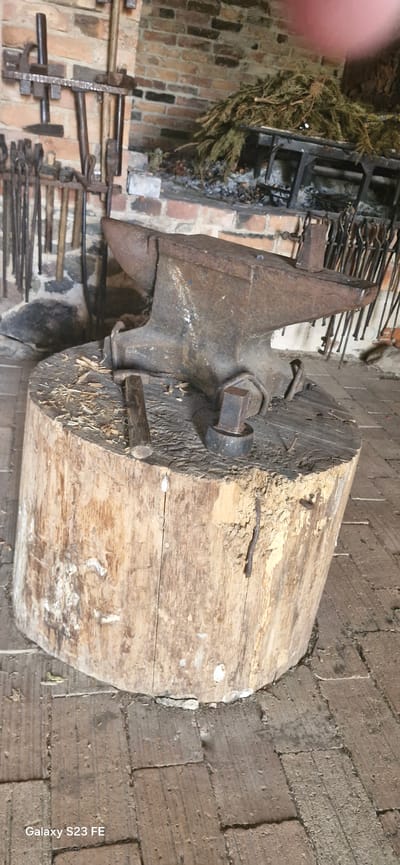 The image depicts a rustic blacksmith's forge at the manor of Pakruojis, Lithuania. In the foreground, a large, weathered anvil rests atop a thick, aged wooden stump. Wood shavings and small pieces of wood are scattered around the base of the stump, suggesting recent use or activity. A small hammer lies on the surface of the stump next to the anvil. Behind the anvil, a brick fireplace is visible, containing a grate with what appears to be dried branches or foliage and ashes, indicating it was recently used for heating. A collection of blacksmithing tools, including tongs and hammers, are mounted on a rack on the brick wall to the left of the fireplace. The floor is paved with brick. The overall scene conveys an authentic historical setting of a working forge.