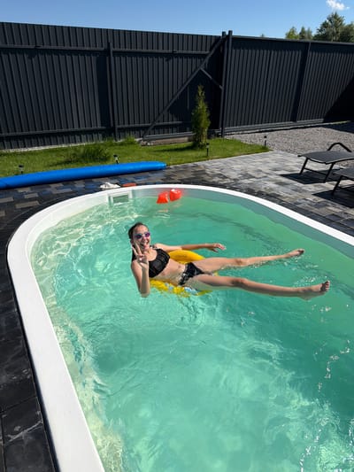 The image shows a young person swimming in an above-ground pool in Sharovechka, Ukraine. The pool is filled with clear, turquoise water and has a white rim. A yellow inflatable ring is partially submerged in the water, and the swimmer is lying on their back with their legs extended upwards. The person is wearing a black bikini and pink swimming goggles, and they are smiling and making a peace sign with their hand. Two small orange inflatable buoys are visible near the person's head. The pool is surrounded by dark paving stones and green grass. A dark corrugated metal fence is visible in the background, with trees and a clear blue sky beyond. A blue rolled-up pool cover lies on the grass next to the pool. The lighting suggests it is daytime, likely a sunny day. The overall mood of the image is one of fun and enjoyment. There is no visible text in the image.