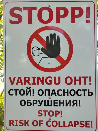 The image shows a white rectangular sign with red and black text and a prohibition symbol. The sign is positioned outdoors, with green foliage visible in the background, suggesting a natural or park-like setting. The sign prominently displays the word "STOPP!" in large red capital letters at the top. Below this, a red circle with a diagonal line across it encloses a black silhouette of a hand, indicating a prohibition.

Further down the sign, there is text in multiple languages. The first line reads "VARINGU OHT!" in red capital letters, which is Estonian for "Danger warning!". Below this, in black capital letters, are Russian words "СТОЙ! ОПАСНОСТЬ ОБРУШЕНИЯ!", translating to "Stop! Danger of collapse!". Finally, at the bottom of the sign, in red capital letters, it says "STOP! RISK OF COLLAPSE.". The presence of these multiple languages suggests the sign is located in a region with diverse linguistic communities or a popular tourist destination. The context provided mentions the city of Ontika, Estonia, which aligns with the Estonian text. The sign itself appears to be made of metal and is mounted with screws, as visible in the image. The overall impression is a warning sign intended to prevent entry or activity due to a potential hazard.