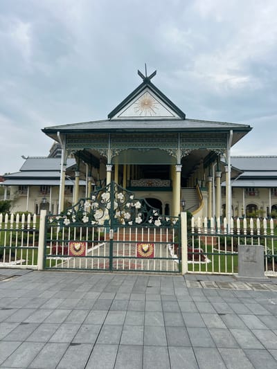 The image shows the main entrance to a grand, colonial-style building, likely a palace or government building, in Alor Setar, Malaysia. The building features a multi-tiered roof with dark grey tiles, a wide veranda supported by numerous columns painted yellow and white, and ornate green and gold decorative elements along the eaves and railing. A prominent triangular gable at the front of the roof displays a sunburst motif within a decorative frame and a crest at the apex.

In front of the building is a decorative green metal gate with intricate scrollwork featuring floral motifs. The gate is flanked by a white picket fence, with the gate and fence section at the entrance to the building having a more substantial and decorative appearance. Behind the fence, a neatly manicured lawn is visible, interspersed with some small red and white flowers and white gravel.

The foreground consists of a grey paved walkway made of large rectangular tiles, with visible seams. The sky is overcast and cloudy, suggesting a dull or perhaps rainy day. No people are visible in the image. The overall impression is one of formal architecture and well-maintained grounds. This building is likely the Istana Anak Bukit, the royal palace of the Sultan of Kedah, located in Alor Setar.