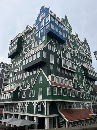 This is a photograph of the Inntel Hotels in Zaandam, Netherlands. The building's facade is designed to resemble a stack of traditional Dutch houses, with a variety of colors including green, blue, and brick red. The architecture features stepped gables and numerous windows, creating a visually complex and striking appearance. The building is tall and appears to be a modern hotel. There is no visible activity or event taking place, and no people are clearly identifiable. The weather appears overcast, contributing to a muted color palette. Visible text on the building includes "Inntel hotels" on a circular sign and "Food & Drinks" on a sign below a tiled roof. The image is oriented vertically, with the building appearing to rise from the bottom of the frame.