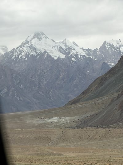 The image shows a panoramic view of a mountainous landscape. In the foreground, there is a vast, barren plain with sparse vegetation. The middle ground features a slope covered in gravel and rocks, leading up to a series of imposing, jagged mountains. The highest peaks are capped with snow, starkly contrasting with the muted tones of the lower slopes. The sky is overcast with grey clouds, suggesting a cool or possibly damp weather. A cluster of small structures, possibly a village or settlement, is visible on the plain, with dust or smoke rising from it. The scene is devoid of any visible people or vehicles, lending it an isolated and remote feel. The image captures the raw, untamed beauty of a high-altitude, mountainous region. The location is specified as Stongde, India.