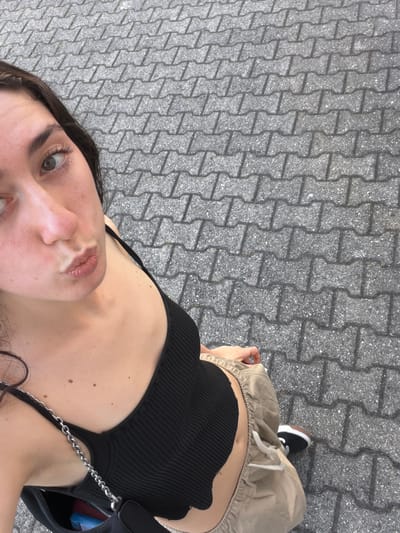 A woman takes a selfie while standing on a gray paved surface with an I-shaped brick pattern. She wears a black ribbed tank top, beige shorts, and a black and white sneaker on her right foot. A black purse with a silver chain strap is partially visible. The background consists entirely of the gray paved surface. The image was taken from a high angle looking down on the subject. The city of Lissone, Italy, cannot be confirmed from the image.
