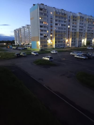 The image captures an exterior view of a residential area in Kopeysk, Russia, at dusk. Towering apartment buildings with numerous windows, some illuminated, dominate the left side of the frame. A paved road runs alongside the buildings, with several cars parked at angles. The road is lined with grassy areas. The sky is a deep blue, indicating the transition from day to night. The scene is quiet, with no visible people or animals. The lighting suggests it is either early morning or late evening. The buildings appear to be modern, multi-story structures typical of urban housing.