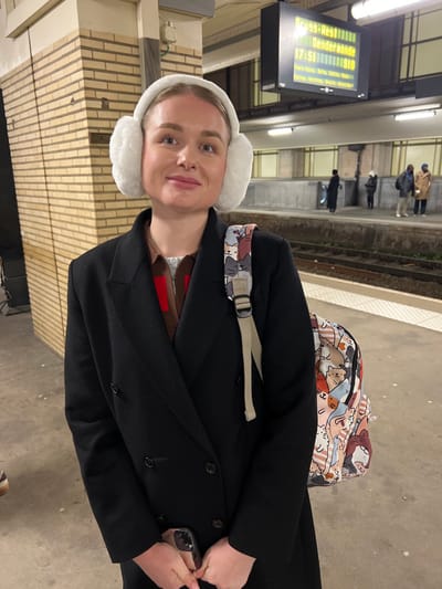 A young woman is prominently featured in the foreground of this image, looking towards the camera with a subtle smile. She wears white, fluffy earmuffs and a dark, long coat over a brown undergarment. A light-colored backpack with a distinctive pattern of various cartoon animals, possibly cats, is visible on her back, with a strap over her right shoulder. She has light-colored hair and a nose piercing, and her hands are clasped in front of her, holding a smartphone with a clear case.
The setting is a dimly lit train or metro station platform, consistent with locations found in Saint-Josse-ten-Noode - Sint-Joost-ten-Node, Belgium. The time of day appears to be late afternoon or early evening, suggested by the artificial lighting and a digital information display in the background. This display clearly shows "17:51" (5:51 PM), along with destination names like "BRUSSEL-RESTRICTIE" (or "Brussel-Restigie") and "DENDERMONDE", and other train details such as "Punct-Tijdig: 20TH. ONTHL. 30TH. ONTHL. MELINE".
In the blurred background, several other individuals are visible standing on the platform, seemingly waiting for public transport. The platform features brick-patterned walls and a concrete ground, with train tracks partially discernible beyond the platform edge. The woman's warm attire, including earmuffs and a coat, suggests cool or cold weather. The image captures a candid moment at a public transportation hub.