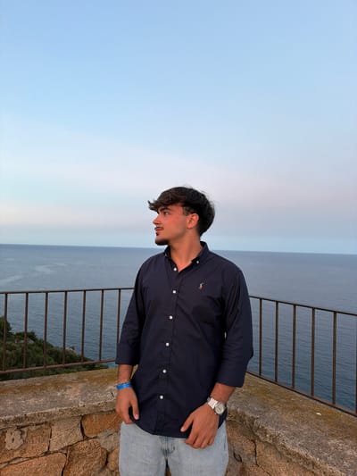 A young man with dark, styled hair and a light beard stands on a stone wall overlooking the sea in Santa Maria Navarrese, Italy. He is wearing a dark blue collared shirt with a small embroidered logo on the left chest and light-wash jeans. A silver watch is visible on his left wrist, and a blue bracelet is worn on the same wrist. The man is looking to his right, away from the camera. Behind him, a metal railing runs horizontally, separating the wall from the sea. The sea is a deep blue, meeting a pale blue and pink sky at the horizon. Green foliage is visible on the cliffside below the railing. The overall impression is a tranquil scene at dusk.