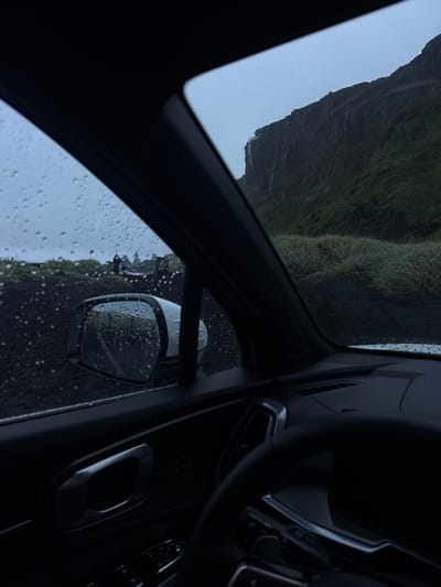 The image captures the view from inside a car on a rainy day in Vik, Iceland. A mountain with a waterfall cascading down its side is visible through the front window. The side mirror, wet with raindrops, reflects a group of tourists and the surrounding landscape. The car's interior elements, including the steering wheel and dashboard, are also in view. The overall atmosphere is dark and overcast due to the rain.