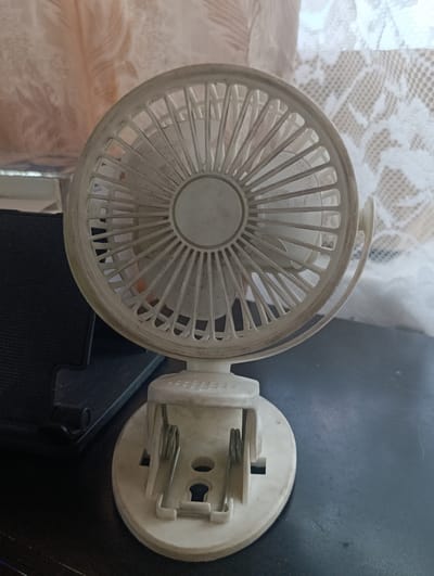 This is a close-up, eye-level shot of a small, white, clip-on fan. The fan is the primary subject, positioned centrally in the frame. It is made of white plastic and appears to be slightly dusty, with visible grime on the blades and casing. The fan is mounted on a white clip base, which is also visibly worn. To the left of the fan, a portion of a black object, possibly a speaker or other electronic device, is visible. The background is softly blurred, featuring a light-colored, textured curtain or fabric with a lace pattern on the right, and a lighter, feathery pattern on the left. The overall setting appears to be indoors, on a dark, possibly wooden surface. There is no indication of time of day, weather, or specific emotions conveyed. No visible text is present in the image. The image is a still photograph, and no activity or event is taking place.