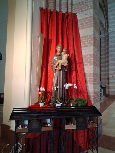 This media file shows a statue of Saint Anthony of Padua holding the infant Jesus. The statue is displayed in a church setting, as evidenced by the brick architecture and the ornate ironwork surrounding the display. A red curtain serves as a backdrop for the statue, and white flowers are placed at its base, suggesting a devotional offering. The lighting appears to be artificial, typical of an indoor environment. The statue is positioned on a raised platform or altar, common for religious figures in churches. To the right of the statue, within the iron enclosure, there are several small objects that appear to be votive candles or offerings. The overall scene evokes a sense of reverence and religious devotion. There is no visible text in the image. The location context suggests this is likely a church in Padua, Italy, a city strongly associated with Saint Anthony. The time of day is not discernible, and the weather is irrelevant given the indoor setting.
