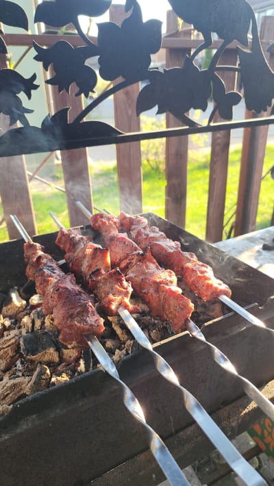 The image depicts a barbecue scene in Рыбхоз, Russia. Several metal skewers, each loaded with chunks of cooked meat, are laid across a charcoal grill. Smoke rises from the meat. In the background, a wooden fence and foliage are visible. A decorative metal screen with a floral design is in the foreground.