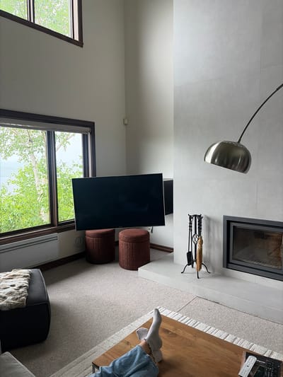 A spacious, modern living room in Mont-Tremblant, Canada, features a tall ceiling with large windows overlooking a lush green landscape and body of water. A large, dark flat-screen television is mounted on the wall, positioned between two round, upholstered ottomans. To the right of the television, a sleek, minimalist fireplace is built into a wall clad in large, light gray tiles. Above the fireplace hangs a modern floor lamp with a metallic dome shade. A set of fireplace tools, including a shovel, poker, and brush, rests on a stand next to the fireplace. In the foreground, a person's legs, clad in blue jeans and white socks, are visible resting on a wooden coffee table. The room is carpeted with a neutral-colored rug.