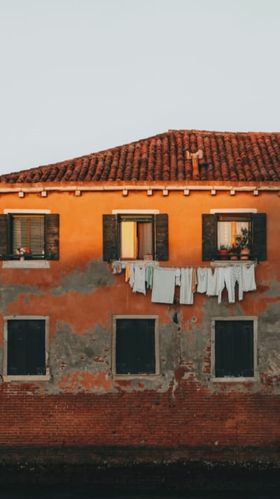 The image displays the exterior of a weathered building, likely an apartment building, with an orange stucco facade that shows signs of age and peeling paint, revealing underlying gray plaster and red brick. The building has a terracotta tile roof and multiple windows. On the second level, there are three windows. The leftmost window has dark wooden shutters, which are partially open, revealing blinds and a vase of flowers on the sill. The middle window, also with dark wooden shutters, is open, showing an interior space and a glimpse of sunlight. Hanging below these two windows, suspended on a clothesline, is a collection of white and light-colored laundry. The rightmost window on this level has dark wooden shutters, and through it, one can see plants in pots on a windowsill. Below these, on the first level, are two more windows with closed dark wooden shutters. The lower portion of the building is predominantly exposed red brick, and the very bottom of the image shows a dark, water-like surface, possibly a canal or river. The sky above is a pale, overcast gray. There are no people visible in the image. The scene suggests a residential building in an urban environment, perhaps a historic district. The provided context specifies that this location is in the city of Oreshki, Russia.