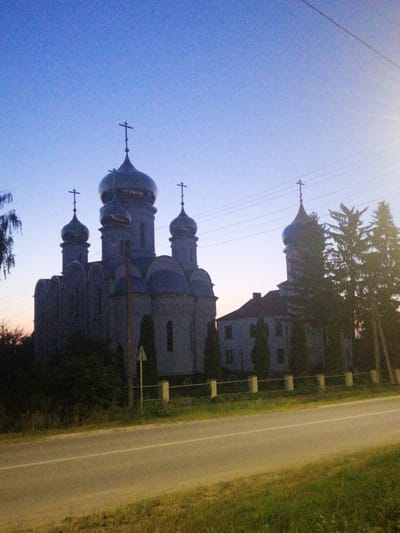 The image shows an Orthodox church with multiple domes and crosses, silhouetted against a twilight sky. The church appears to be a large, imposing structure with a complex architectural design. Several trees, including what appear to be cypress trees, surround the church. A road runs in front of the church, with a grassy verge on either side. Power lines are visible stretching across the sky. The overall impression is one of a serene and somewhat somber scene, perhaps captured at dusk or dawn. The image is of the Transfiguration Cathedral in Lanivtsi, Ukraine.