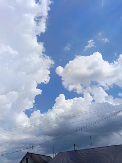 A low-angle, vertical shot captures a bright blue sky filled with dramatic, puffy white cumulus clouds. In the foreground, the dark, corrugated metal roofs of two houses are visible, with one house featuring a peaked roof and a window. Power lines crisscross horizontally across the sky above the houses, and an antenna with multiple radiating arms is mounted on the roof of the house on the right. A faint plume from a chimney is also visible on the roof of the house on the left. The scene suggests an outdoor environment in the city of Toguchin, Russia, under a partly cloudy sky.