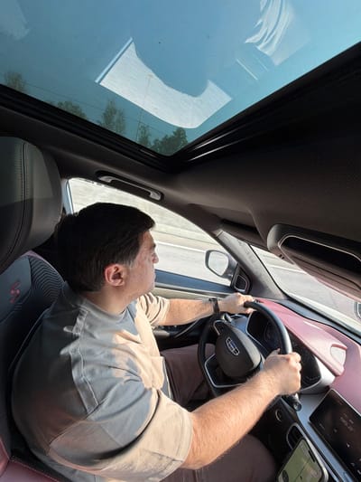 In the city of Fyodorovo, Russia, a man is driving a car with a panoramic sunroof open. He is wearing a light-colored t-shirt and brown pants. The interior of the car is visible, including the steering wheel, dashboard, and seats. The sunroof allows a view of the sky and trees.
