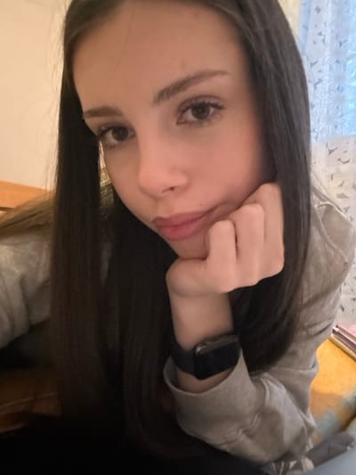 The scene is an indoor, close-up portrait of a single individual, located in the city of Волховец, Russia. The subject is a young adult female with long, straight dark brown hair, brown eyes, and fair skin. She is looking directly towards the camera with a neutral expression, her lips slightly pursed. Her left hand is raised, with fingers curled, supporting her chin. She is wearing a light grey, long-sleeved garment, possibly a sweatshirt. On her left wrist, a dark-colored smartwatch with a dark band is visible.
The immediate background on the left side of the frame features a light brown wooden surface, possibly part of furniture. On the right side, a white or light-colored curtain with a floral or intricate pattern is visible, obscuring a window. Below the curtain and to the right of the subject's arm, blurred objects are discernible, including a pink-covered item resembling a book or notebook and a light green or blue object. The lighting appears soft and diffused, characteristic of an indoor setting with ambient light sources.