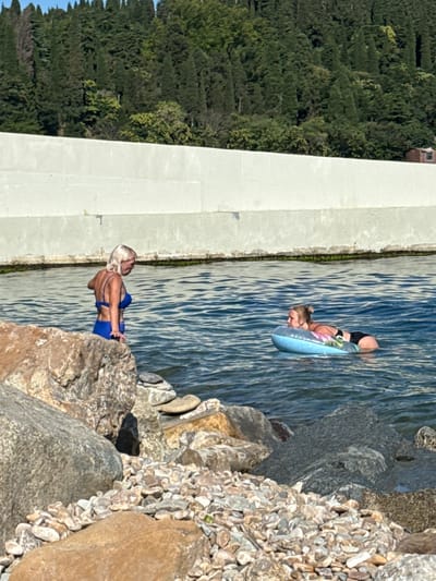 Two women are in the water near a rocky shore. The woman on the left, with blonde hair, is standing in the water wearing a blue bikini. The woman on the right is lying on a blue inflatable ring, wearing black shorts and a dark-colored top. In the background, there is a concrete wall and a dense forest on a hillside. The sky is clear and blue. The location is Ut'os, Ukraine.