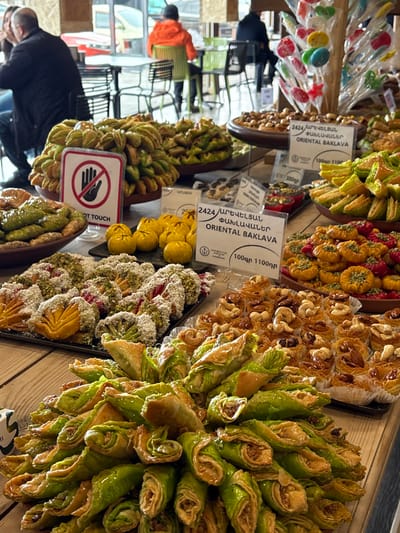 This image depicts the interior of a confectionery or bakery in Tsovagyugh, Armenia, showcasing an extensive array of oriental sweets and baklava. The immediate foreground is dominated by a large pile of green-hued, rolled baklava pieces, likely pistachio-filled, resting on a wooden surface. To the left, a bowl of similar green pastries is visible. Further into the scene, multiple trays and platters present a variety of sweets, including golden-yellow, rounded pastries; sweets topped with green pistachio crumbs, white coconut, and red elements; and numerous individual, noodle-like pastries crowned with whole nuts such as cashews and almonds.
Several white informational signs are interspersed among the displays, prominently featuring "2424 / ԱՐԵՎԵԼՅԱՆ ՓԱԽԼԱՎԱՆԵՐ / ORIENTAL BAKLAVA" in both Armenian and English, accompanied by pricing (e.g., "100gr 1100դր"). A red-and-white circular sign displaying a white hand with a diagonal line across it, indicating "DO NOT TOUCH," is positioned in front of a tray of green pastries.
In the blurred background, several patrons are seated at light-colored tables with a mix of brown and lime green chairs. A man in a dark jacket is partially visible on the far left, while another person in a bright orange jacket is seated with their back to the camera. Further to the right, a person in a dark jacket is also seated. Behind the seating area, large windows reveal an indistinct outdoor street scene, including a yellow vehicle. A decorative stand with colorful lollipops is visible on the right side of the background display. The overall environment suggests a clean and well-lit commercial establishment with light-colored walls and flooring.