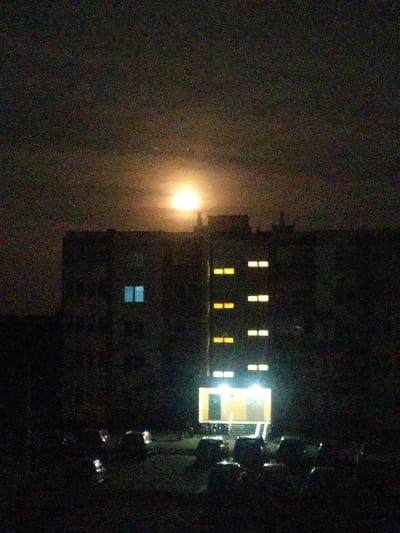 This image captures a night scene in the city of Velikalukski rural Council, Belarus. A large, multi-story building dominates the frame, with several lit windows visible on its facade, emitting a warm yellow glow. Two windows on the left side of the building are lit with a cooler blue light. At the base of the building, a brightly lit entrance with stairs is visible. In front of the building, a dark foreground reveals the silhouettes of parked cars, suggesting a residential or apartment complex. The sky above is dark and hazy, with a prominent, luminous full moon shining brightly, its light diffused by the atmospheric conditions. The overall impression is one of a quiet, nocturnal urban landscape illuminated by artificial lights and the natural light of the moon.