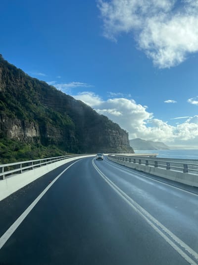 The image displays the Sea Cliff Bridge, located within the Wollongong City Council area, Australia. A two-lane asphalt road, dark and reflective, curves to the right from the foreground. White double solid lines mark the road's center, and a single solid white line borders the left shoulder. To the immediate left, a massive, steep rock cliff face, densely covered with green vegetation, rises vertically alongside the road. A light-colored concrete barrier topped with a silver guardrail separates the road from the cliff. To the right, an identical concrete barrier and silver guardrail delineate the road from the blue waters of the Pacific Ocean, where small waves or whitecaps are visible. In the distance, across the ocean, multiple layers of hazy green and blue mountains or headlands are discernible. One white SUV-type vehicle is observed traveling in the distance on the curved road, moving away from the viewer. The upper portion of the scene is dominated by a clear blue sky with scattered, bright white cumulus clouds. The environment is naturally lit during daylight hours.