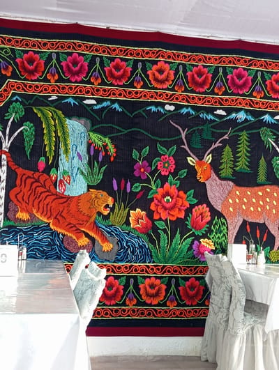 The image depicts a brightly colored, embroidered tapestry hanging on a wall in what appears to be a dining establishment in Bosteri, Kyrgyzstan. The tapestry features a vibrant scene with a tiger in the foreground, leaping near a waterfall and a body of water. To the right, a deer with antlers stands amidst stylized trees and flowers. The tapestry is bordered by ornate floral patterns. In front of the tapestry, two tables are set with white tablecloths, and several chairs with white slipcovers are visible. Salt and pepper shakers and a table number sign are present on one of the tables. The environment suggests an indoor setting, likely a restaurant or cafe, with the tapestry serving as a prominent decorative element. There are no people clearly visible in the image.