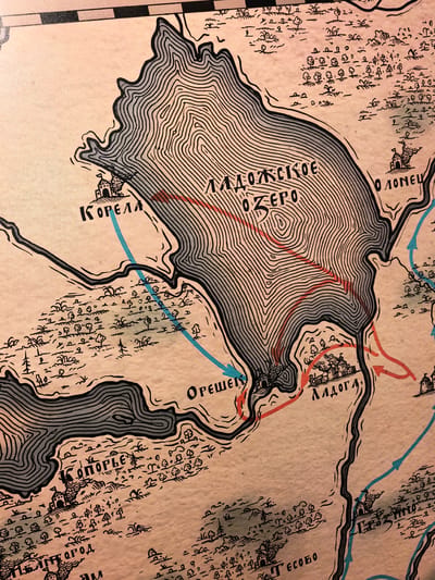 The image is a section of an old, stylized map of Staraya Ladoga, Russia. The map depicts a large body of water, labeled "ЛАДОЖСКОЕ ОЗЕРО" (Ladoga Lake), with contour lines indicating elevation. Several settlements are marked with fortified structures, such as "КОРЕЛА" (Korela), "ОРЕШЕК" (Oreshek), "ЛАДОГА" (Ladoga), and a partially visible "ППГОРOД" (Ppgorod) and "ЕСОБО" (Esobo). Red and blue arrows are drawn on the map, suggesting routes or movements, possibly military. The map features small drawings of trees, grass, and other vegetation. The overall aesthetic is that of an antique illustration with a limited color palette of black, grey, and beige, with red and blue accents for the arrows.