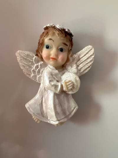 This is a close-up, eye-level, indoor studio shot of a small angel figurine, likely made of resin, against a plain, neutral-toned background. The figurine depicts a baby angel with light brown curly hair adorned with a white floral halo. It has large, wide-open blue eyes, rosy cheeks, and a small pink mouth. The angel is wearing a flowing white gown with a high neckline and is holding a light-colored sphere or ball in its hands. Its wings are spread outwards, with detailed feather textures. The lighting is soft, casting a subtle shadow to the right of the figurine. There is no visible indication of its location within the city of Constanța, Romania; it is presented in an isolated, generic setting.