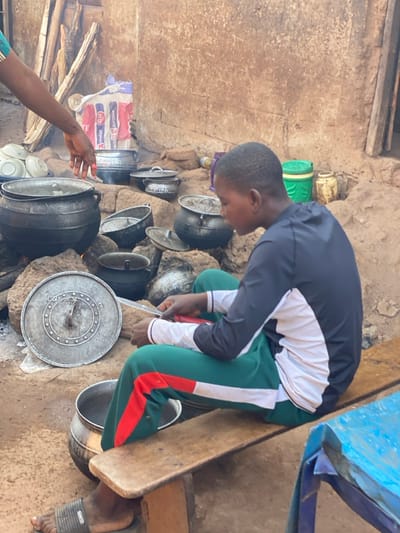 A person kneels beside an earthen wall adorned with cooking pots of varying sizes. The individual wears a black jacket with white sleeves and green pants with a red stripe. They appear to be stirring or preparing food in one of the pots. To the left, another person's arm reaches toward the cooking area near a bag that is leaning against the wall. A blue plastic chair sits partially visible in the upper right corner. The scene gives an impression of daily life around cooking.