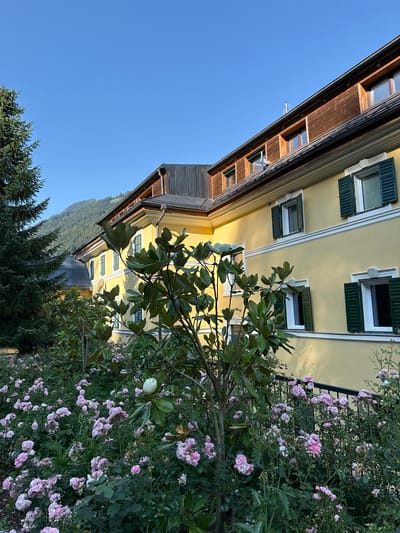 In Bad Gastein, Austria, a large, yellow, two-story building with a dark grey roof and wooden trim is bathed in sunlight. The building features multiple windows with dark green shutters, some of which are open. In front of the building, a lush garden is filled with blooming pink roses and various green plants, including a magnolia tree with a prominent white bud. A tall evergreen tree stands to the left of the building, and a wooded mountain can be seen in the background under a clear blue sky.