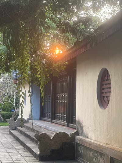 The image captures a section of traditional architecture in Kim Long Ward, Vietnam, under direct sunlight with a visible lens flare emanating from dense green foliage. A prominent light-colored building wall on the right features a circular opening containing a reddish-brown wooden lattice window. The wall's lower section exhibits weathering and discoloration. To the left, a recessed entrance is visible, composed of dark, grid-patterned double doors, accessed by three stone steps. These steps are flanked by ornate, stylized carved stone balustrade elements. Further left, another section of a light-colored wall with blue accents is partially obscured by foliage, revealing a "WC" sign on a white background mounted on a post. The ground leading to the steps is paved with square or rectangular stone blocks. Abundant green foliage, including trailing vines, frames the top and left side of the scene, partially obscuring the rooflines and the sunlight source. A tree with bare branches is visible in the distant left background. No individuals or discernible activities are present.